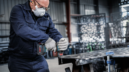 Person i skyddsutrustning använder en elektrisk skruvdragare på metallram i industrimiljö.