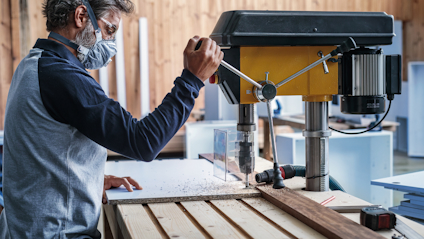 Person använder en gul borrmaskin för att bearbeta trä, bär skyddsmask och glasögon.