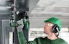 Person i grönt skyddsutrustning använder en grön elektrisk borr på en metallkonstruktion.