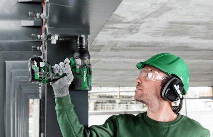 Person i grönt skyddsutrustning använder en grön elektrisk borr på en metallkonstruktion.