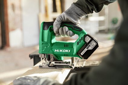 Grön elektrisk sticksåg med märket "HiKOKI", hålls av en hand i arbetshandske över träplanka.