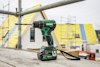 Sladdlös borrmaskin med gröna detaljer placerad på en byggnadsställning. Bostadshus under konstruktion i bakgrunden.