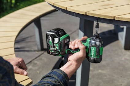 En person håller i en grön och svart sladdlös skruvdragare vid ett träbord utomhus.