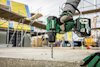 Hand med grön borrmaskin borrar i betongplatta, byggställning och material i bakgrunden.
