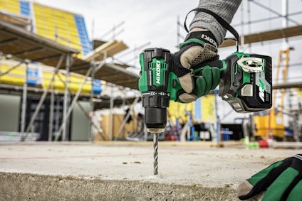 Hand med grön borrmaskin borrar i betongplatta, byggställning och material i bakgrunden.