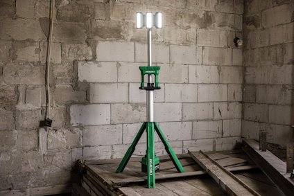 Grön belysningsstativ med vit lampa står i ett rum med grova betongväggar och träplankor på golvet.