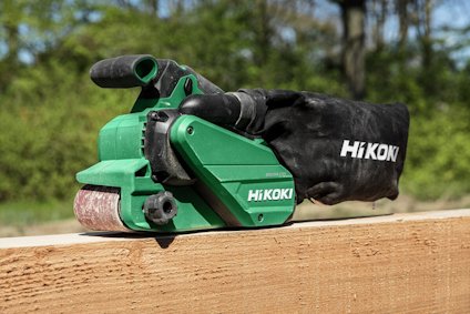 Grön bandslipmaskin av märket HiKOKI används utomhus, placerad på en träbalk.