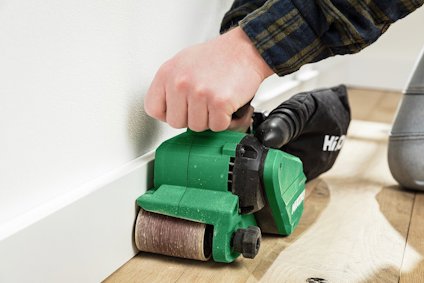 Hand använder en grön bandslip på trägolv nära en vit golvlist.