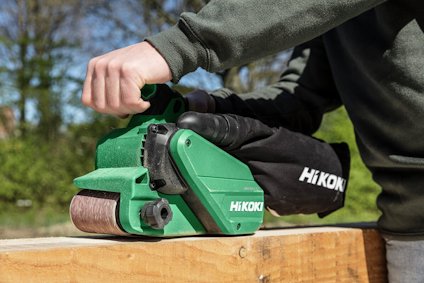 Person som använder en grön HiKOKI bandslipmaskin på en träyta utomhus.
