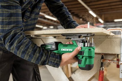 En person använder en grön Hitachi-verktyg för att kapa trä vid en arbetsbänk i en verkstad.