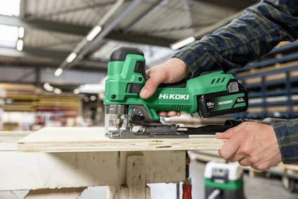Person använder en grön Hikoki sticksåg för att såga i en träskiva i en verkstadsmiljö.