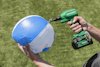 En person blåser upp en blå och vit strandboll med en grön Hikoki pump på en gräsyta.