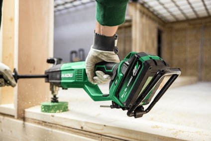 Person använder en grön batteridriven Hitachi-verktyg för att såga trä i en byggnadsmiljö.