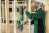 Man with safety gear using a green and black Hitachi power saw on wooden beams.