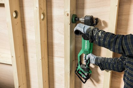 Person använder en grön handhållen borrmaskin mot en trävägg med vertikala reglar.