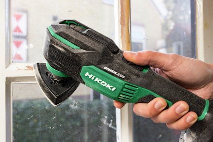 Hand håller i en grön och svart verktyg med texten "HIKOKI" och "BRUSHLESS" nära ett fönster.