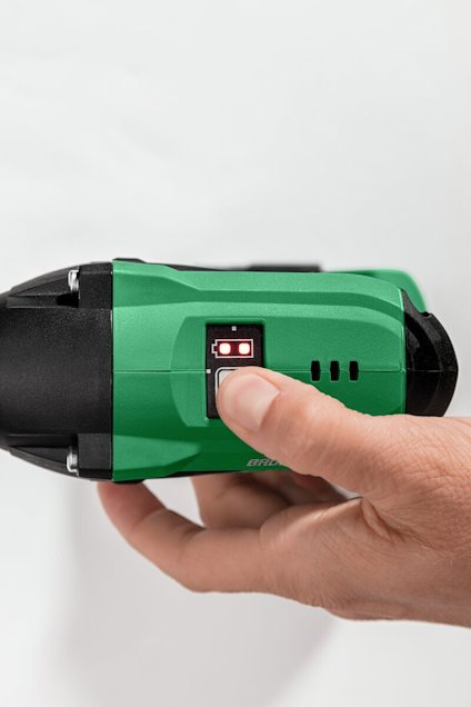En hand trycker på knappen på en grön och svart elektrisk enhet, troligen en borrmaskin.
