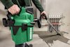 Person bär en grön Hikoki industridammsugare, riktad mot ett vattenläckage på golv nära rör i väggen.
