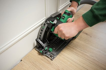 Person använder en cirkelsåg för att skära i trä nära en vit vägg. Sawdust avlägsnas genom en ansluten slang.
