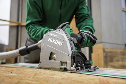 Person i grön tröja använder en grå och grön Hikoki cirkelsåg med ansluten dammsugarslang på en träplanka.