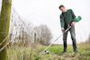 Person som använder en grön trimmer för att klippa gräs vid ett stängsel på en äng.