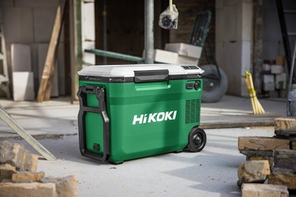 Grön och grå HiKOKI-kylbox med hjul, placerad på ett byggarbetsplatsgolv nära staplade tegelstenar.