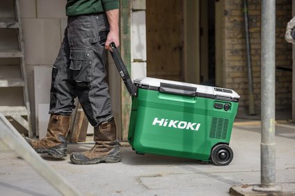 En person i arbetskläder drar en grön portabel kylbox på hjul med texten "HiKOKI" på en byggarbetsplats.