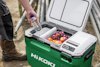 Grön kylbox med öppen lock visar flaskvatten och frukt inuti. Person i arbetskläder står intill boxen.
