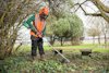 Person i skyddsutrustning använder en röjsåg för att klippa vegetation nära ett träd och bänkar.