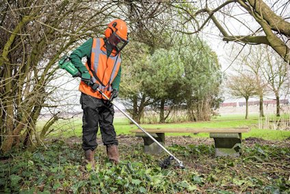 Person i skyddsutrustning använder en röjsåg för att klippa vegetation nära ett träd och bänkar.