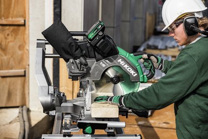 Person i hjälm använder en grön Hitachi kap- och gersåg för att såga en träbit.