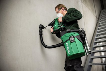Person i skyddsglasögon och munskydd använder en grön och svart HIKOKI borrmaskin mot en betongvägg.