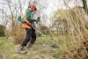 Person med skyddsutrustning använder en grästrimmer på en gräsbevuxen yta med buskar och träd.