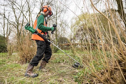 Person med skyddsutrustning använder en grästrimmer på en gräsbevuxen yta med buskar och träd.