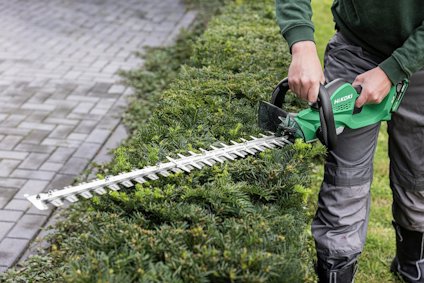 Person i gröna kläder använder en grön Häcksax av märket Hikoki på en tät häck bredvid en stenlagd gång.