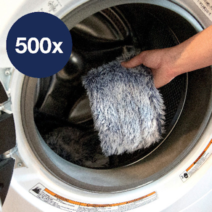 Hand placerar en grå, lurvig trasa i en tvättmaskin. Texten "500x" syns på en blå cirkel i bilden.