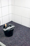 En svart hink med en spackelspade står på en filt vid ett hörn av vitt kaklat vägg.