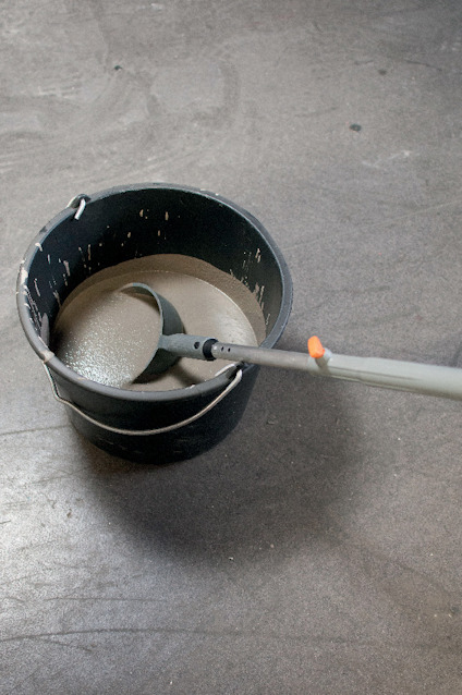 En hink med grå cementblandning och en vispstav nedsänkt i blandningen på grått golv.