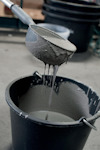En grå hink fylld med flytande cement och en grå slev där cementen droppar ner i hinken.
