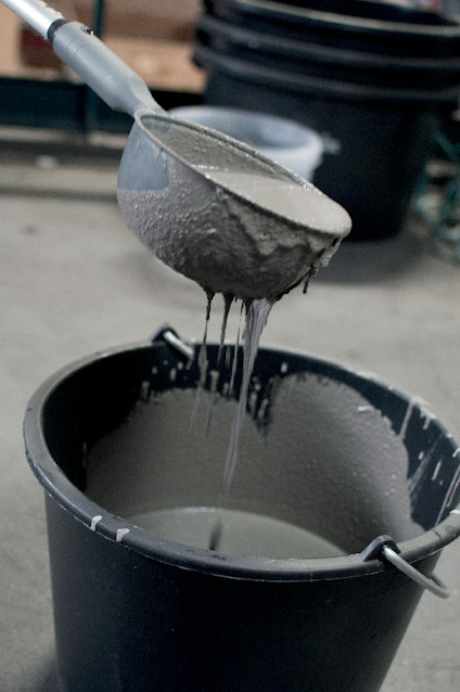 En grå hink fylld med flytande cement och en grå slev där cementen droppar ner i hinken.