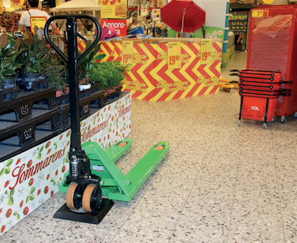 Grön handtruck på butiksgolv, omgiven av skyltar med erbjudanden och staplade varukorgar.