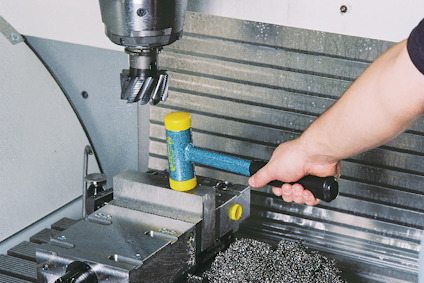 En hand som håller en blå och gul hammare nära en borrmaskin med metallspån på en arbetsbänk.