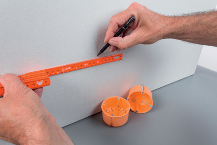En person markerar en punkt på en grå vägg med en orange mätsticka och penna. Två orange installationsboxar ligger bredvid.