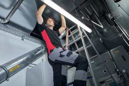 Person i arbetskläder arbetar med belysning i industriellt utrymme. Stege och ventilationsrör syns i bakgrunden.