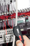 En hand håller en avmantlare som används för att ta bort isolering från en kabel framför ett kopplingsskåp med röda kablar.