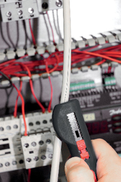 En hand håller en avmantlare som används för att ta bort isolering från en kabel framför ett kopplingsskåp med röda kablar.