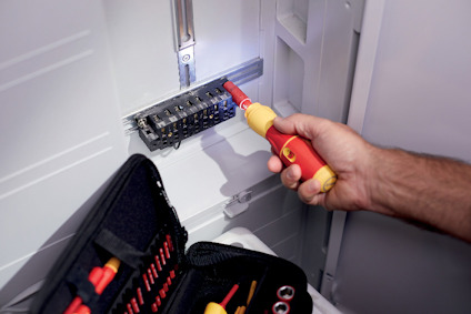 En person använder en testpenna för att justera en elektrisk terminal i ett skåp, verktygssats synlig i förgrunden.