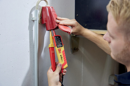 Person använder en röd och gul multimeter för att mäta kabeln som sitter fast på väggen.