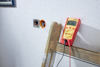 Multimeter visar 0.138 på trästege, bredvid en vägguttag med kablar synliga.