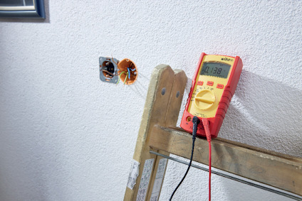 Multimeter visar 0.138 på trästege, bredvid en vägguttag med kablar synliga.